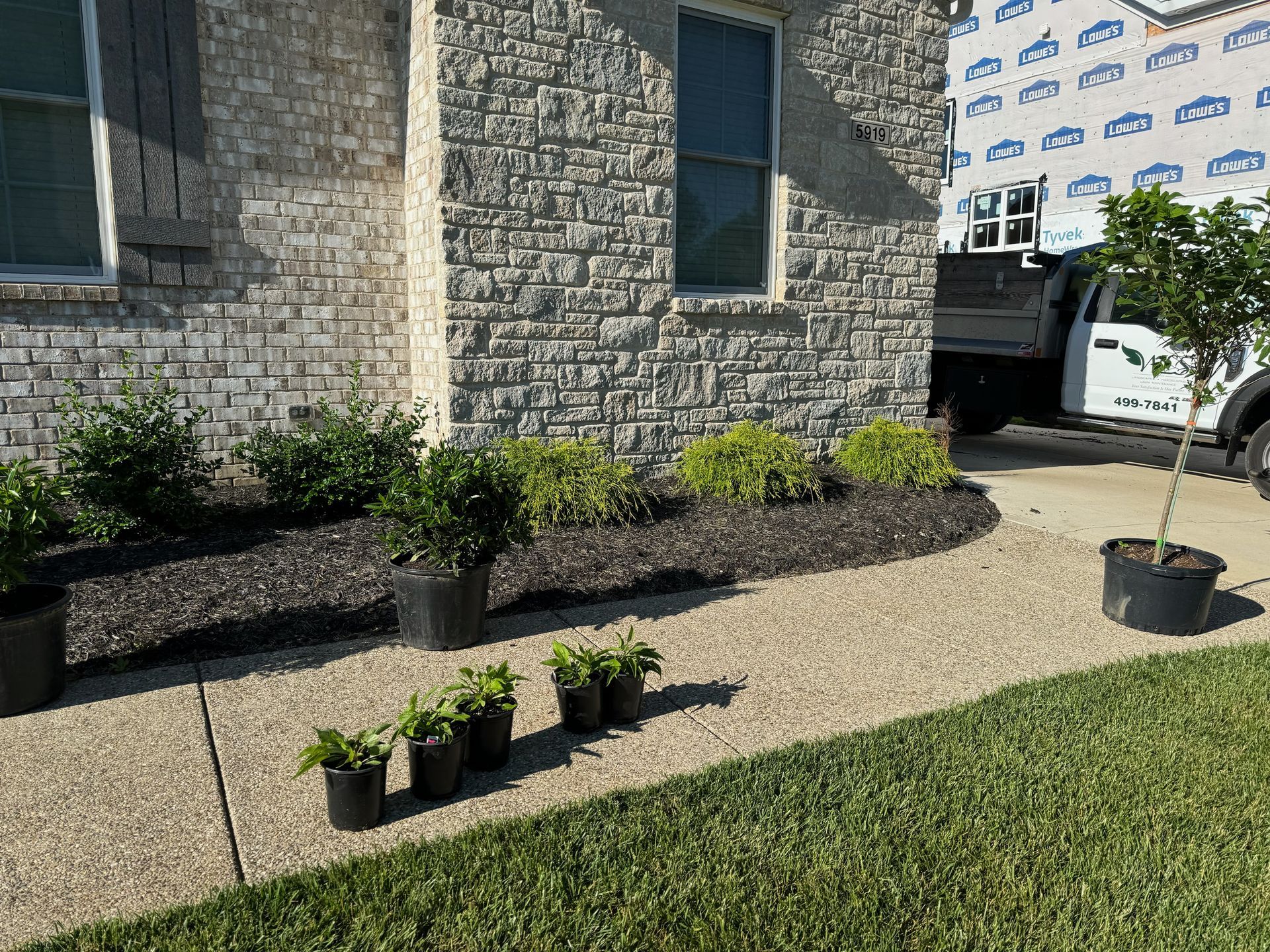 A house with stone veneer, a concrete pathway, and a flower bed. Plants in black pots are lined up along the pathway.
