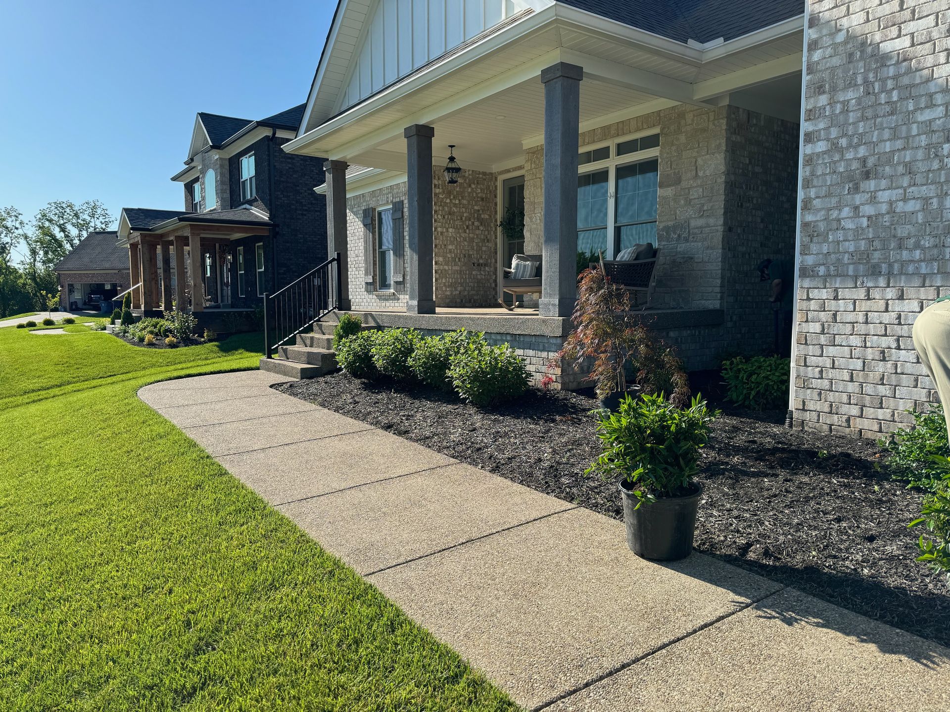 A paved walkway leads to a modern house with gray stone and black mulch landscaping. Green lawn and a blue sky are visible.