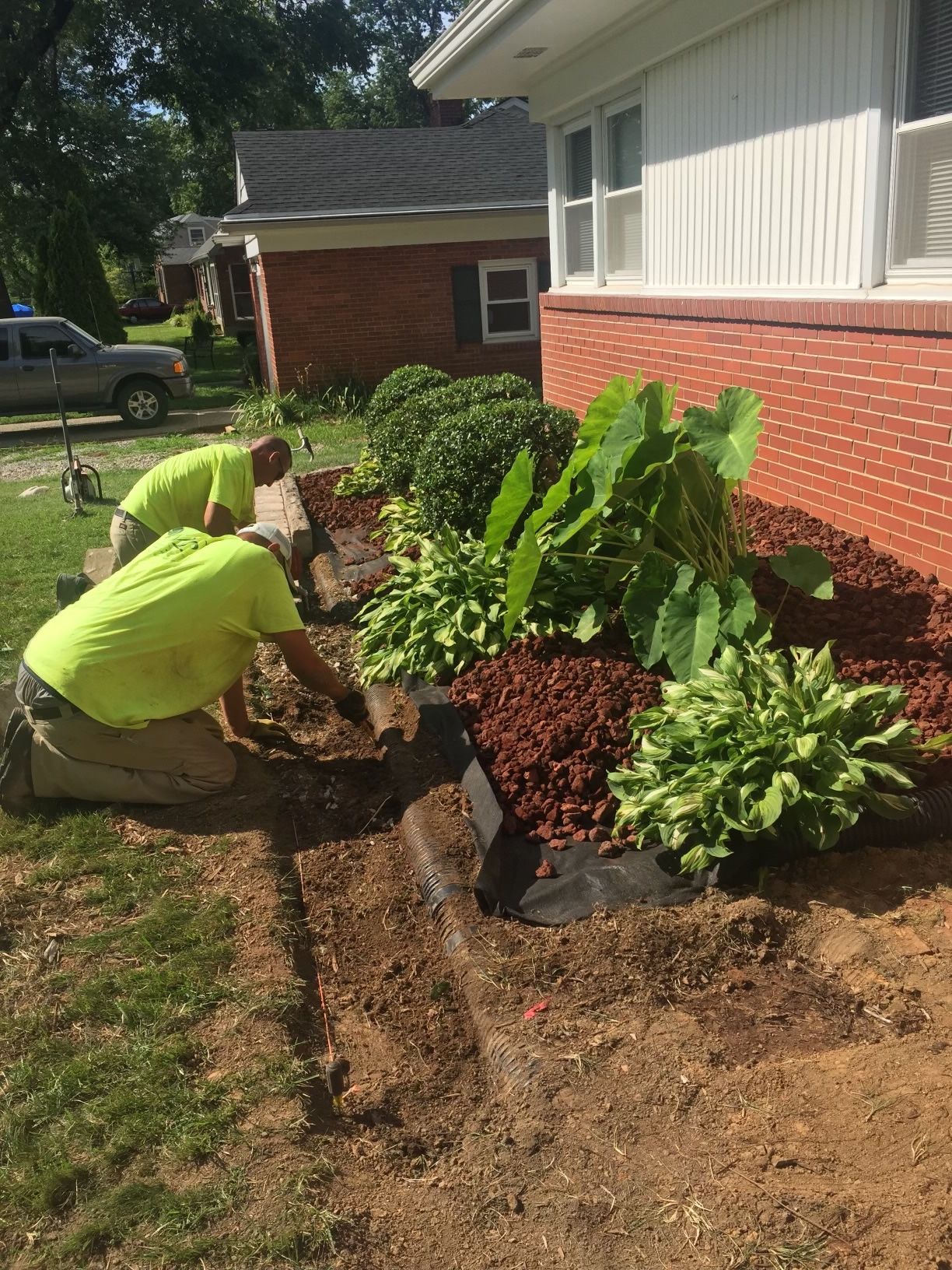 Three workers in neon shirts landscaping a flower bed in front of a red brick house.