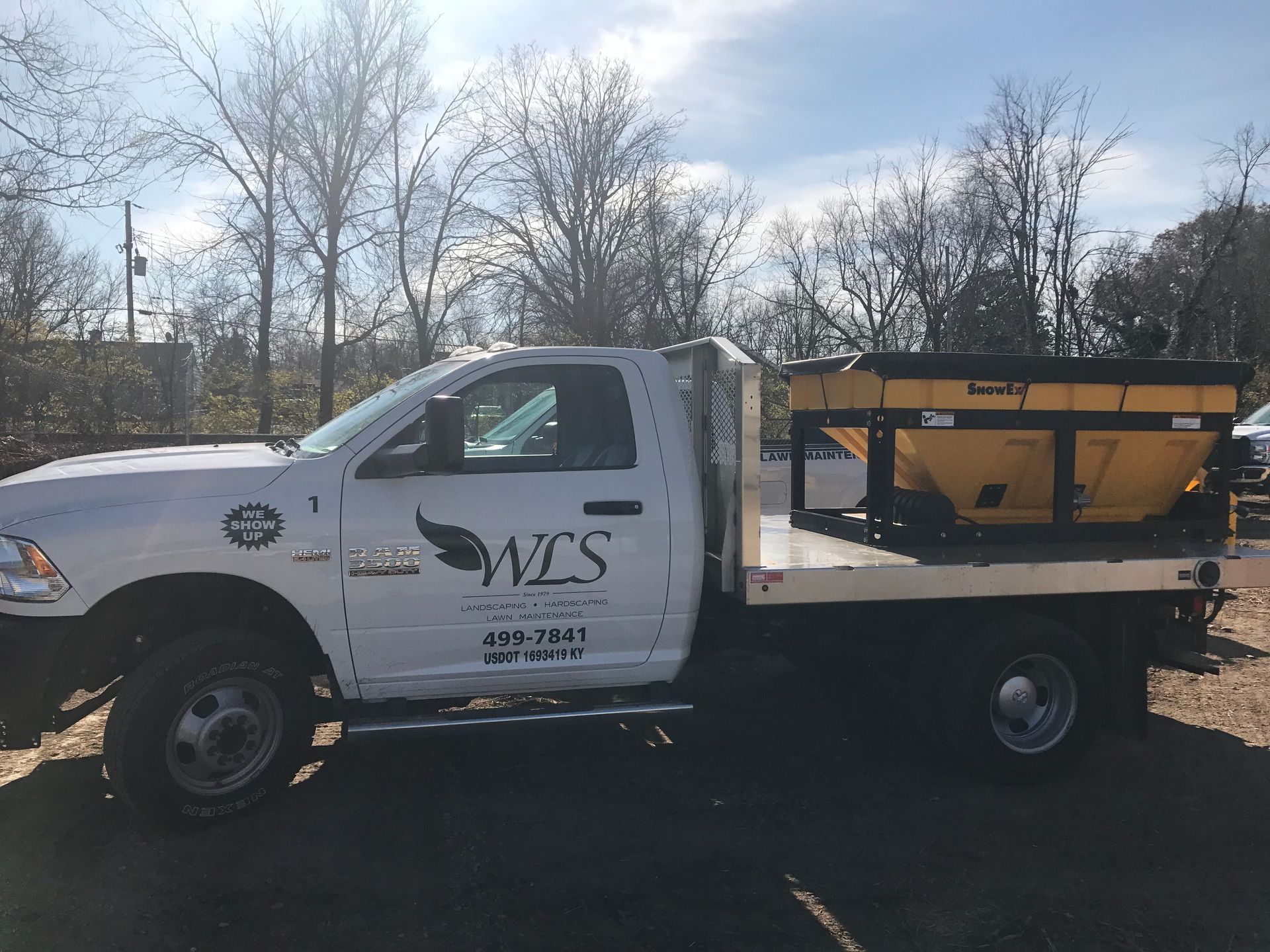 White utility truck with MVS logo carrying a yellow plow attachment on a flatbed in a roadside lot