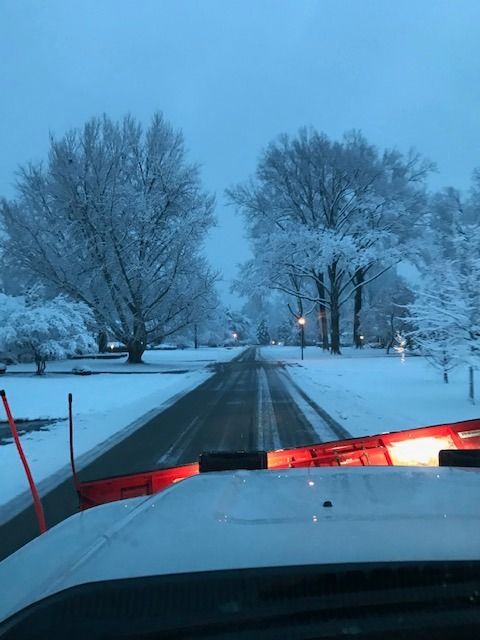 Snowy road at dawn viewed from a plow truck, with red lights and frosted trees lining the street