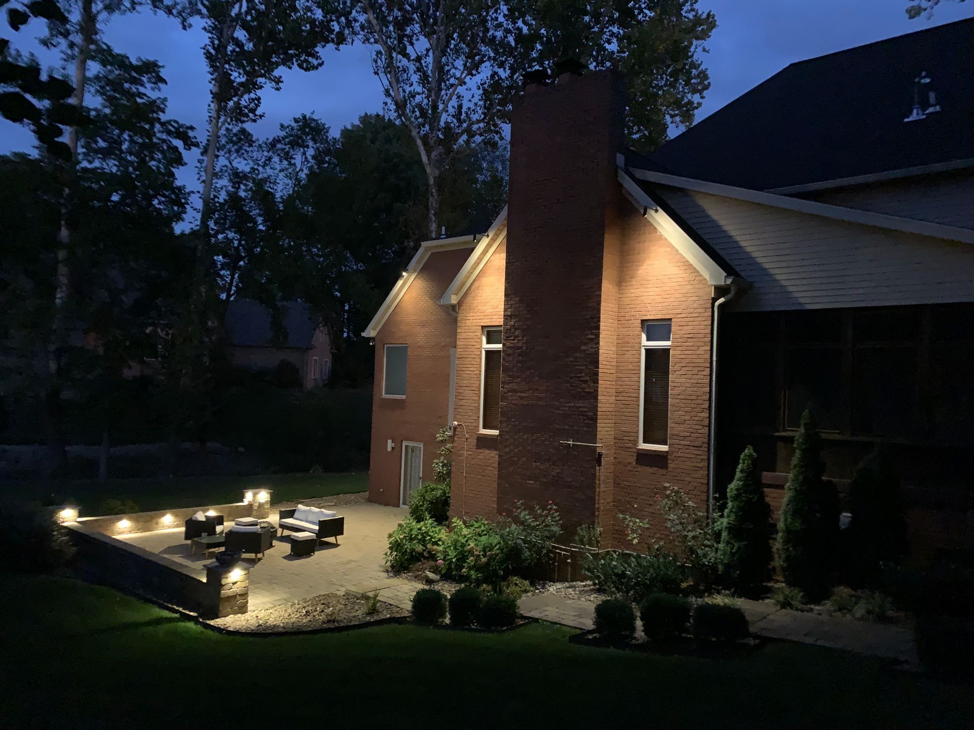 Backyard patio at dusk with string lights, a fire pit, and uplighting on the brick home.