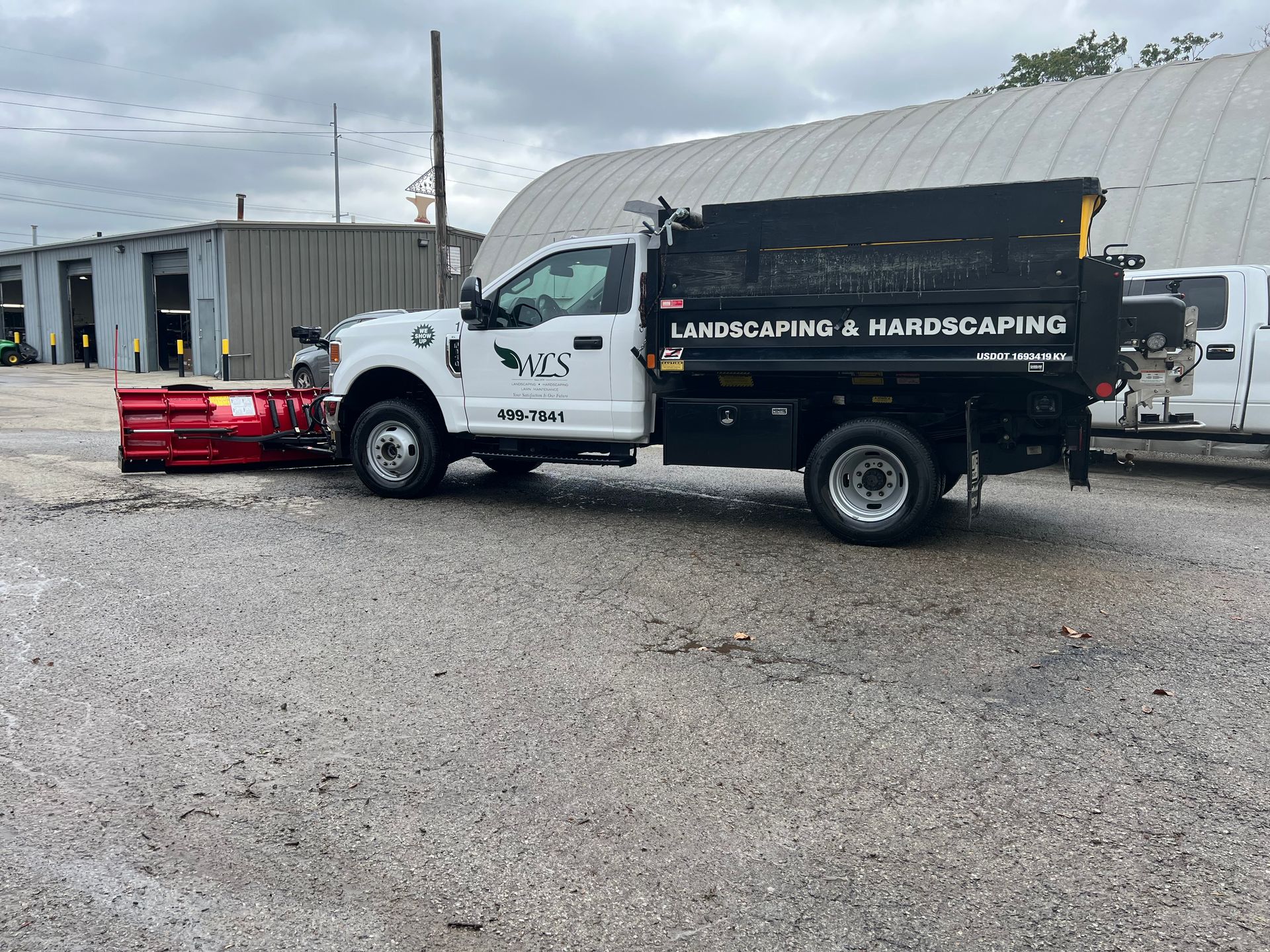 White work truck with black dump bed and red trailer parked on a gravel lot near industrial buildings.