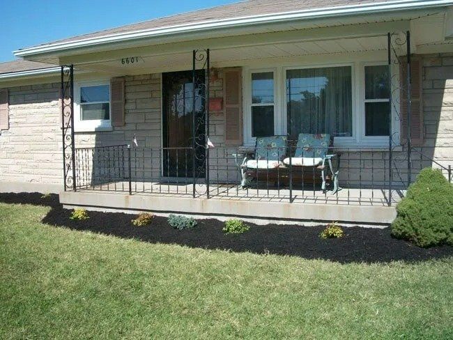 A brick house with a small porch and black railing. A bench sits on the porch. The yard has mulch and green grass.