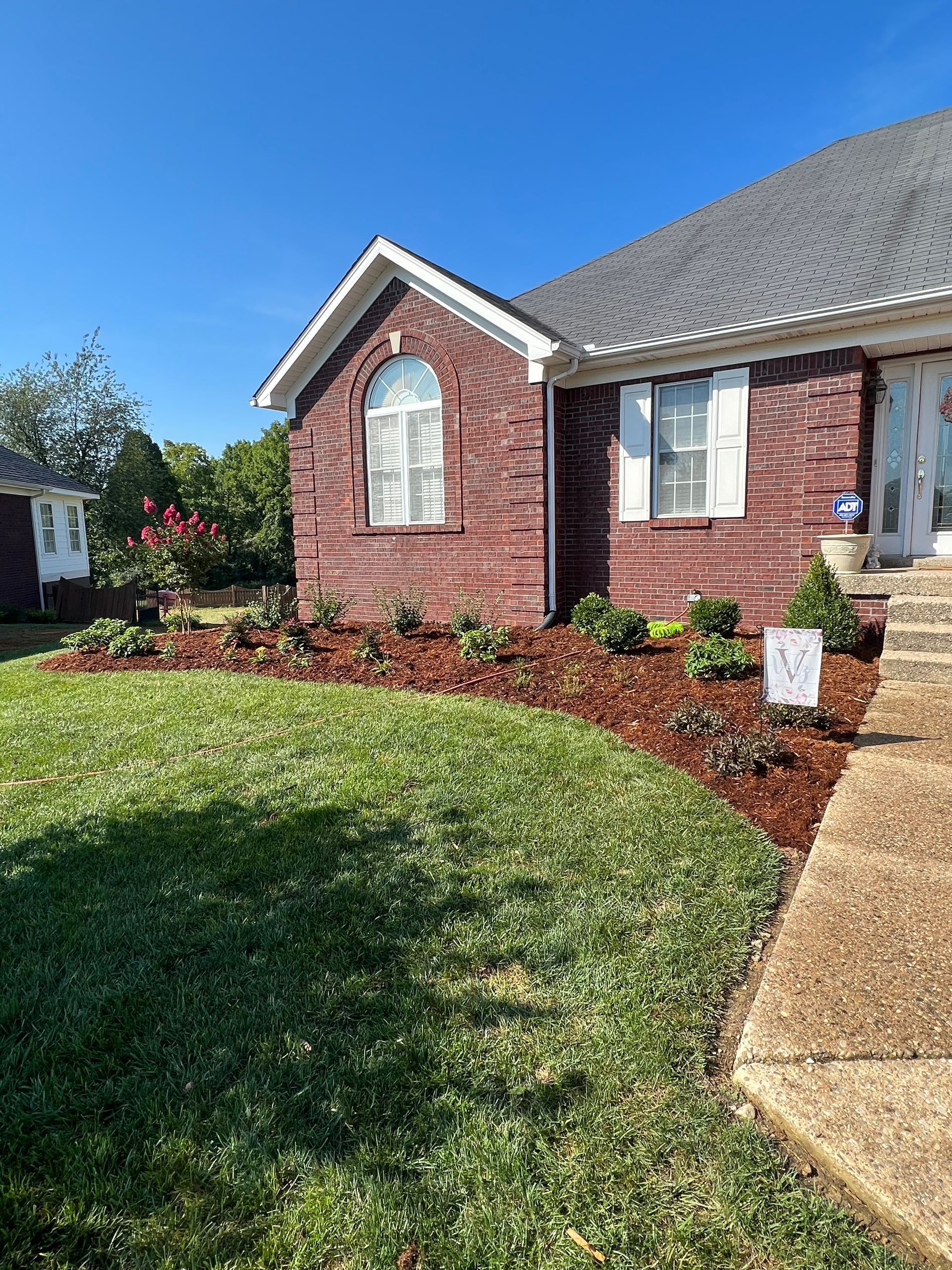 A brick house with a manicured lawn and a flower bed with brown mulch. The sky is blue and sunny.