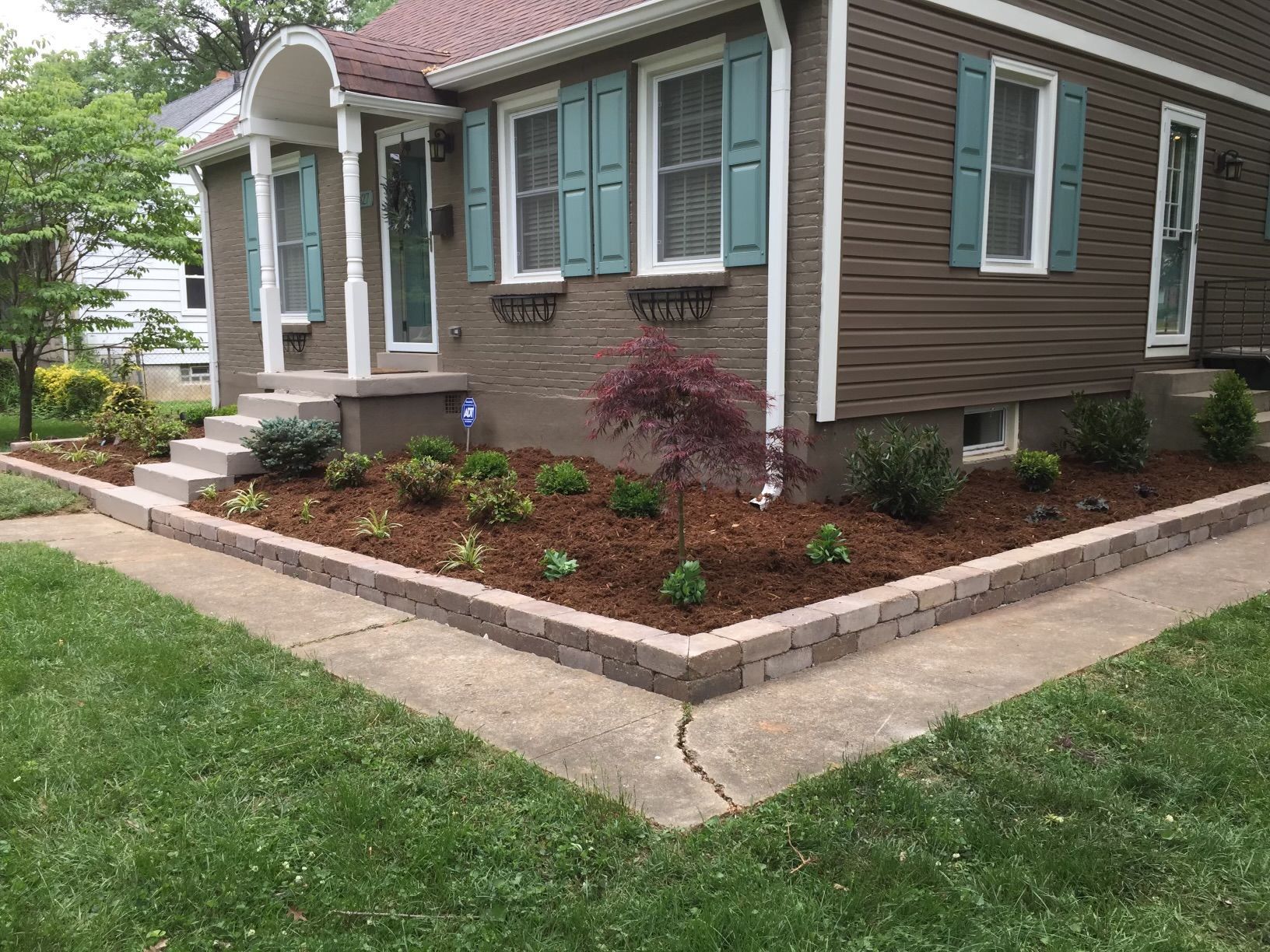 Brown house with teal shutters, featuring a freshly mulched garden bed edged with brick pavers and a concrete walkway.