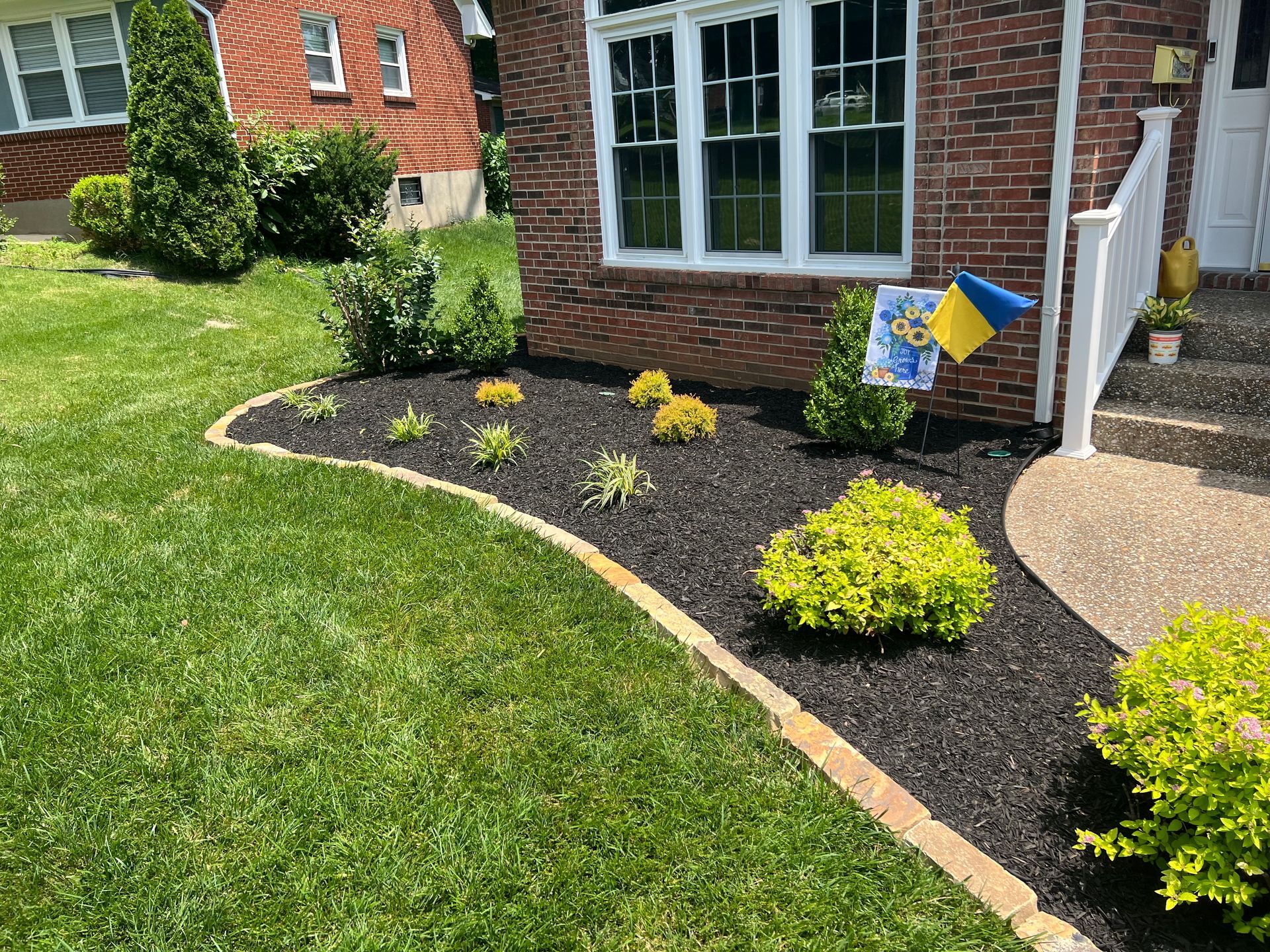A well-manicured front yard with a flower bed edged in stone and filled with black mulch and various green and yellow plants.