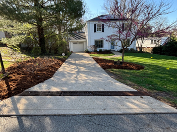 Driveway leading to a white house with a garage, drainage grate, and landscaping with mulch.