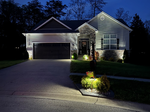 A house at night, illuminated by outdoor lighting on the facade and landscaping, with a driveway leading up.