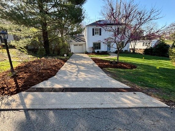Driveway leading to a white house with a garage. A drainage grate spans the width of the concrete driveway.