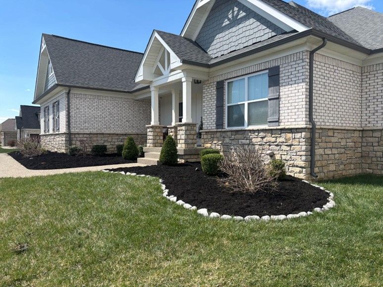 A modern house with a light brick facade, dark gray roof, and a well-manicured lawn with a black mulch bed in the foreground.