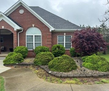 A brick house with a landscaped front yard featuring green bushes and a red Japanese maple tree.