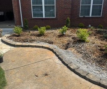 A landscaped flower bed with a stone border in front of a brick house. The bed contains small plants and mulch.