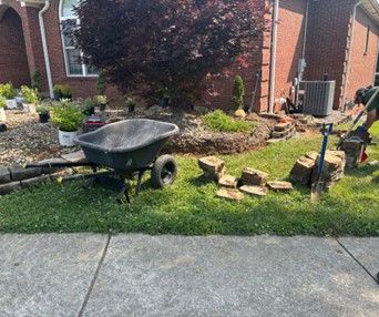 A person working on landscaping in front of a brick house, with a wheelbarrow, rocks, and plants visible.