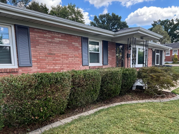 A brick house with blue shutters, a small front porch, and neatly trimmed hedges lining the front yard under a blue sky.