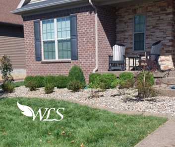 A brick home with a landscaped front yard featuring green bushes, gravel, and a small lawn. Two chairs sit on the porch.