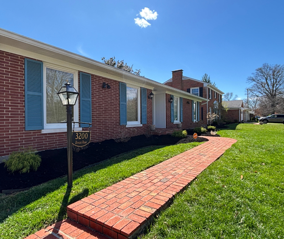 Brick ranch house with blue shutters, a brick walkway, and a manicured lawn under a blue sky.