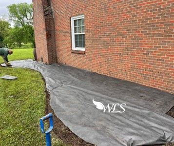 A person lays landscape fabric along the side of a brick building. The yard is green with a blue shovel.