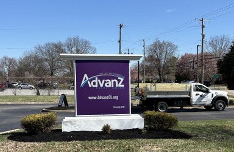 Sign for Advanz, purple background, white lettering, beside a road, with a white truck.