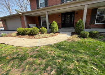A brick house with a front porch, concrete walkway, and landscaped shrubs in front of a green lawn under a sunny sky.