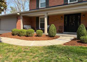 Red brick house with a front porch, garage, and landscaped flower bed featuring small shrubs and mulch.