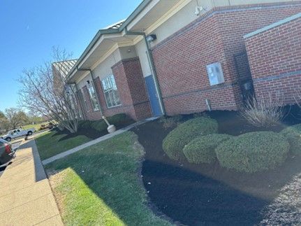 Exterior of a brick building with a paved walkway, green grass, and trimmed bushes on a sunny day.