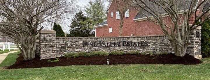 Sign for Pine Valley Estates made of stone and with text in black. Bare trees on either side. Brick building in background.