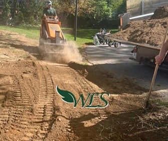 A person operating an orange mini excavator digs a trench in a yard with soil. Someone else holds a rake.