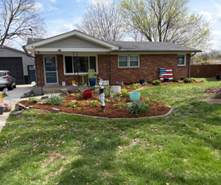 A single-story brick house with a front porch, a mulched garden bed with potted plants, and a lawn under a blue sky. A single-story brick house with a front porch, a mulched garden bed with potted plants, and a lawn under a blue sky.