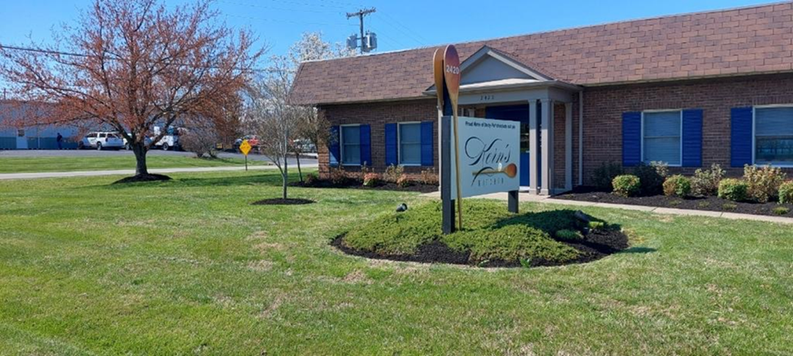 A building with blue shutters and a sign in front, surrounded by a grassy lawn and trees under a clear sky.