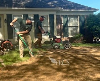 Two men landscaping a yard in front of a house; one rakes the ground, the other uses a machine, under sunlight.