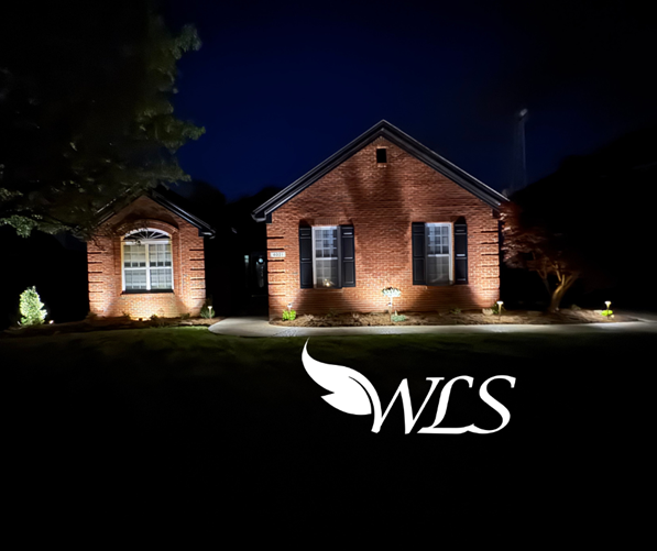 A brick house and small outbuilding are illuminated at night by landscape lighting. The WLS logo appears in the foreground.
