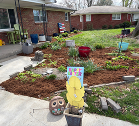 A front yard garden with brown mulch, small plants, a red pot, and an Easter sign near a brick house. A front yard garden with brown mulch, small plants, a red pot, and an Easter sign near a brick house.