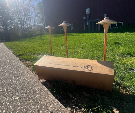 Three bronze landscape lights and a cardboard box on a lawn next to a concrete sidewalk.