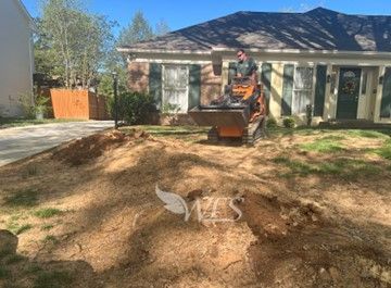 A person operating a mini excavator removing tree stumps in front of a house. The lawn is covered in wood chips.