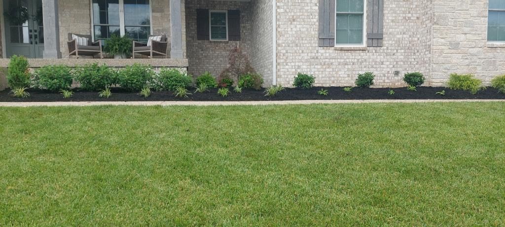 A green lawn in front of a house with a porch and flower bed, the foundation is gray brick.