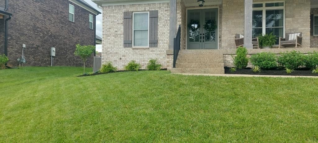 Lawn in front of a brick house with a porch and bushes along the foundation. A second brick house is to the left.