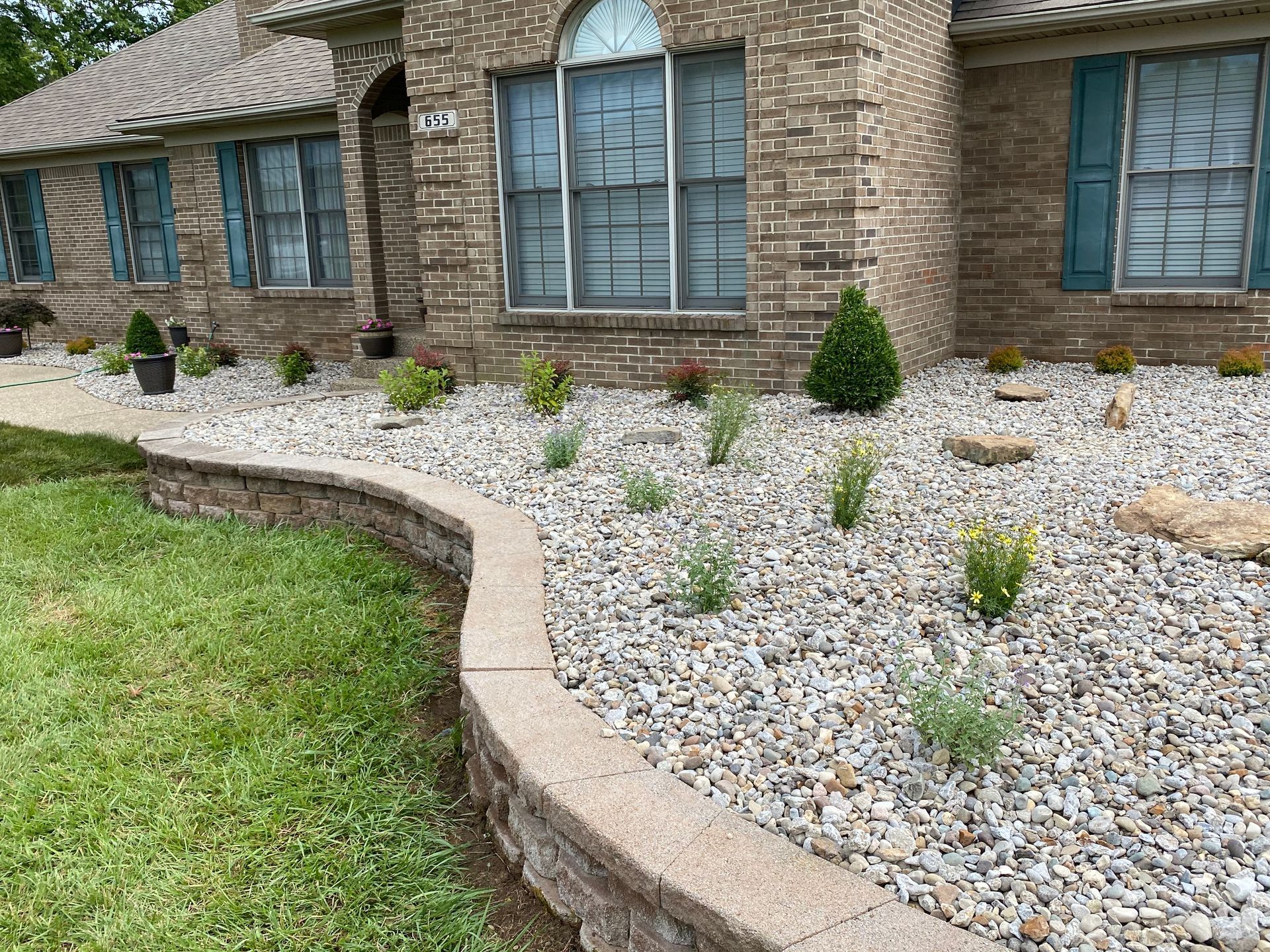A house with a brick facade and a rock garden lined with a low stone retaining wall. Green grass sits to the left.