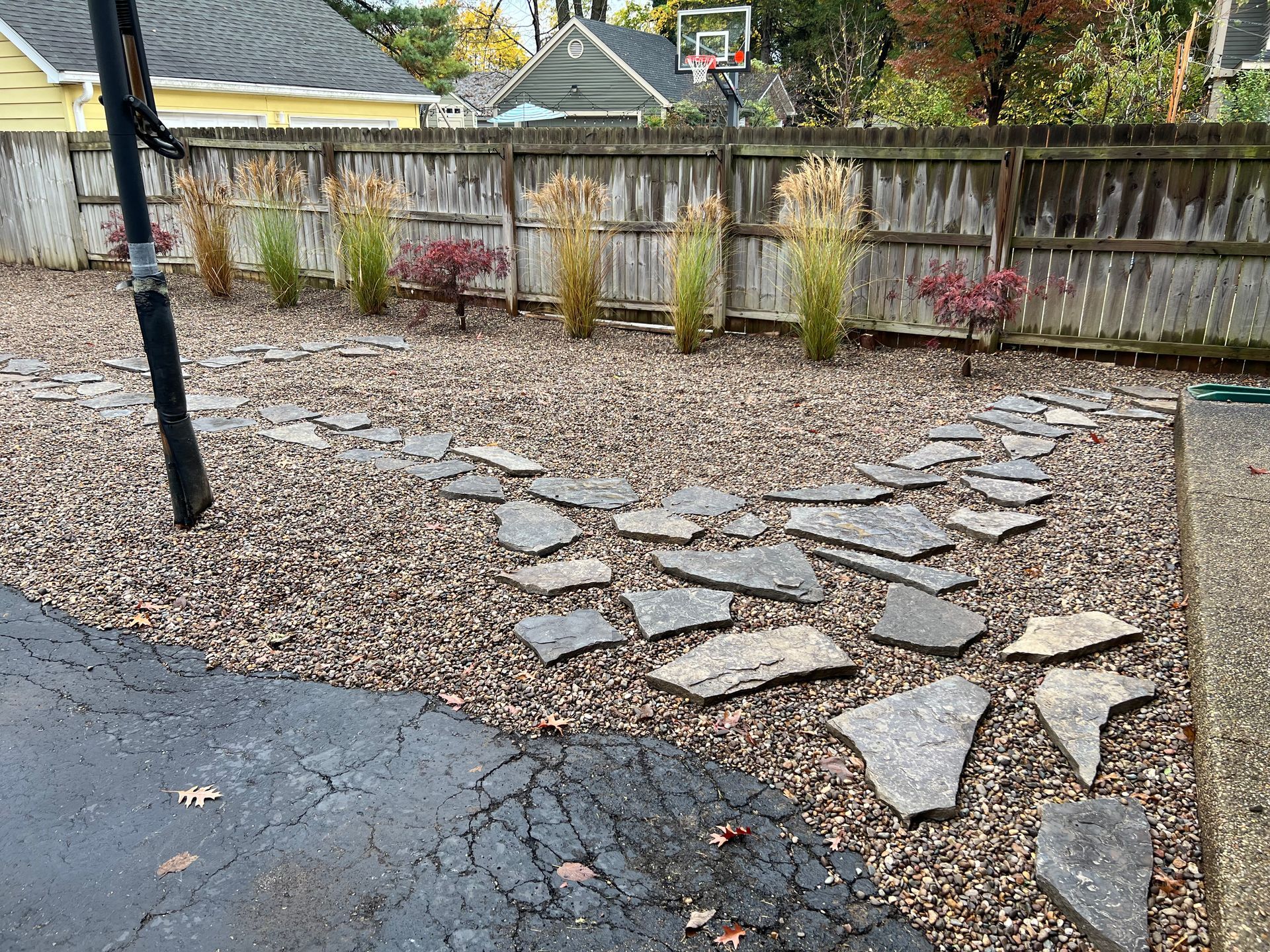 A backyard with a flagstone path and decorative plants in front of a weathered wooden fence. The ground is covered in small brown stones.