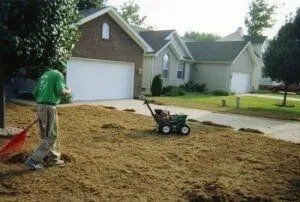 Person spreading soil in a suburban front yard with a wheelbarrow near a driveway and garage