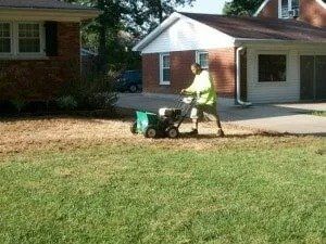 Person using a snow blower on a suburban driveway beside a brick house.