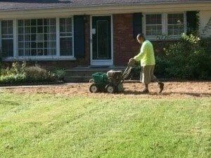 Person mowing a lawn in front of a house with a push mower.