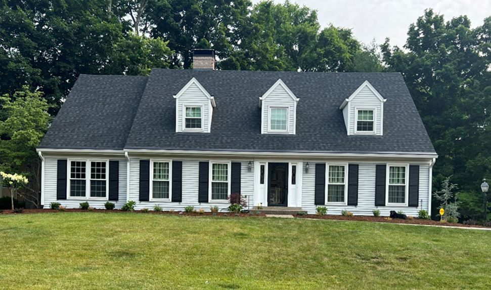 A white house with black shutters, three dormers, and a dark roof sits on a green lawn, surrounded by trees.