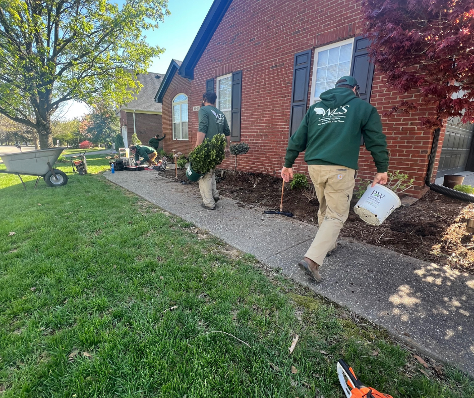 Landscapers in green hoodies and khaki pants work on a flower bed next to a brick house. One carries a bucket, another carries a bush.