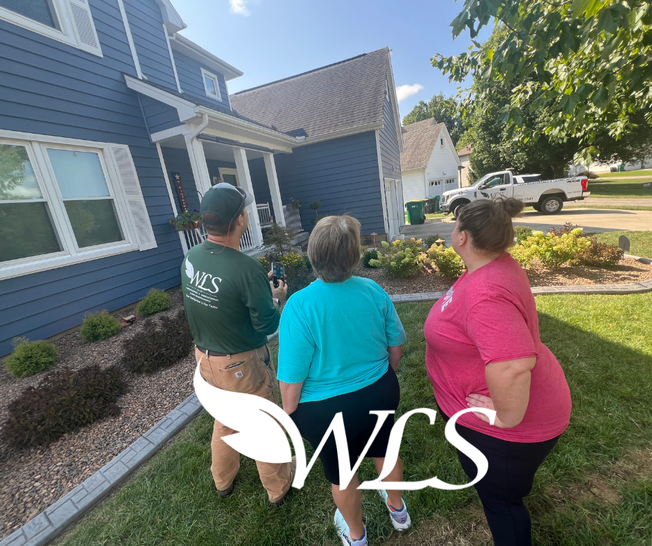 Three people, two women and one man, standing in front of a blue house, presumably discussing landscaping. The man wears a green shirt with 