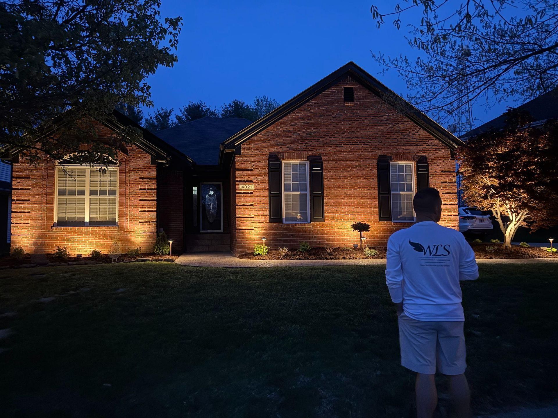 A man stands in front of a brick house illuminated by landscape lighting at dusk.