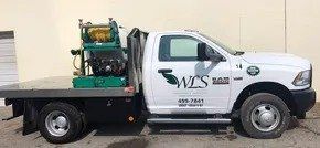 White WCS RAM truck with a flatbed carrying equipment. The truck is parked outdoors against a beige background.