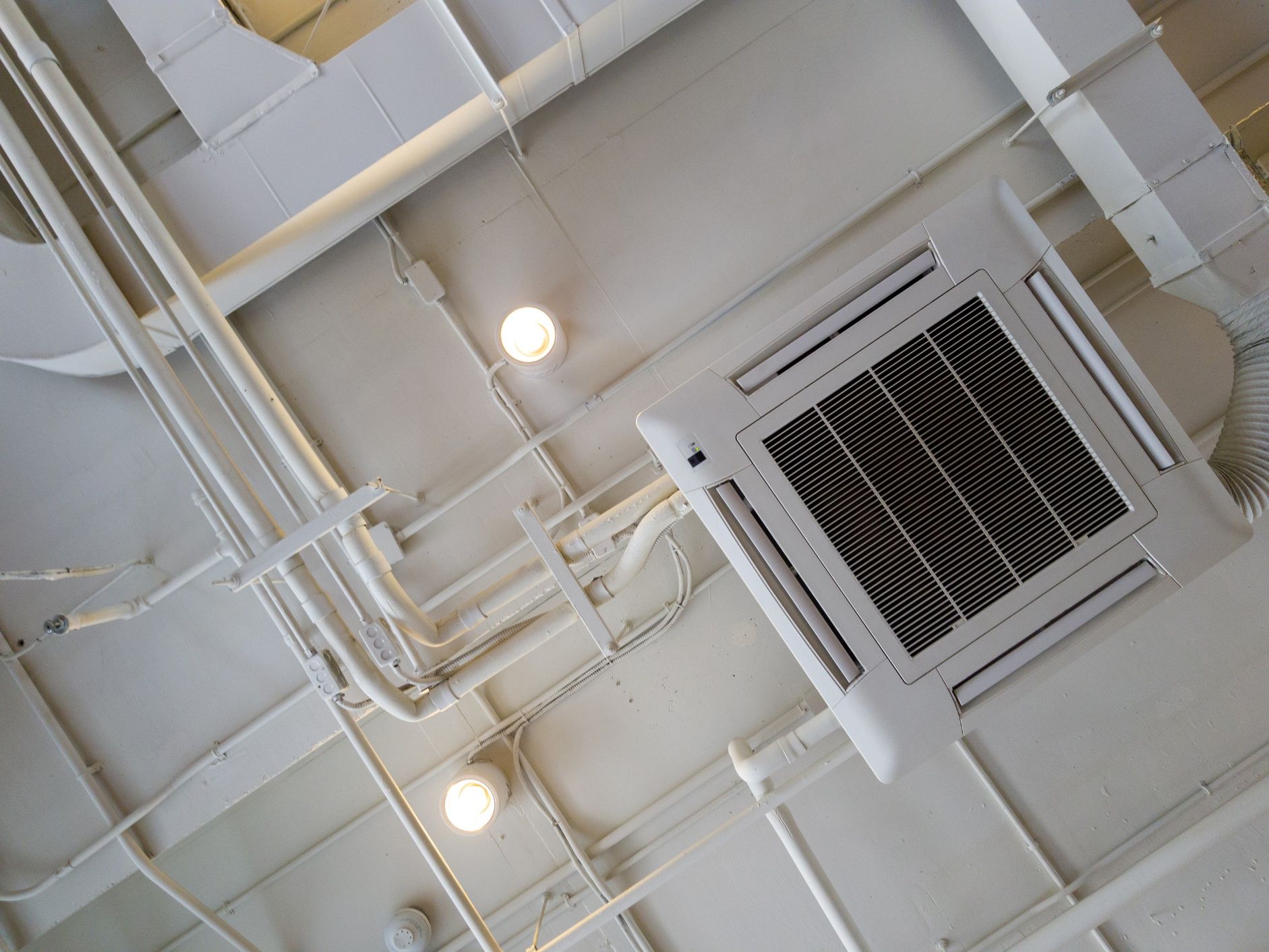 White ceiling with exposed pipes, electrical conduits, air conditioning unit, and circular lights.