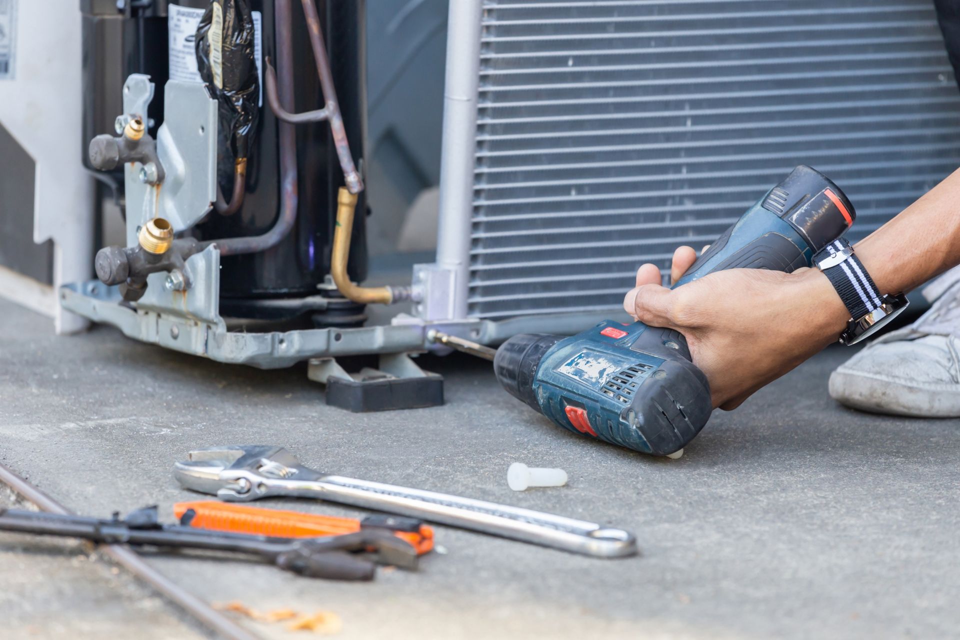 Person using a drill to work on an air conditioning unit; tools on concrete.