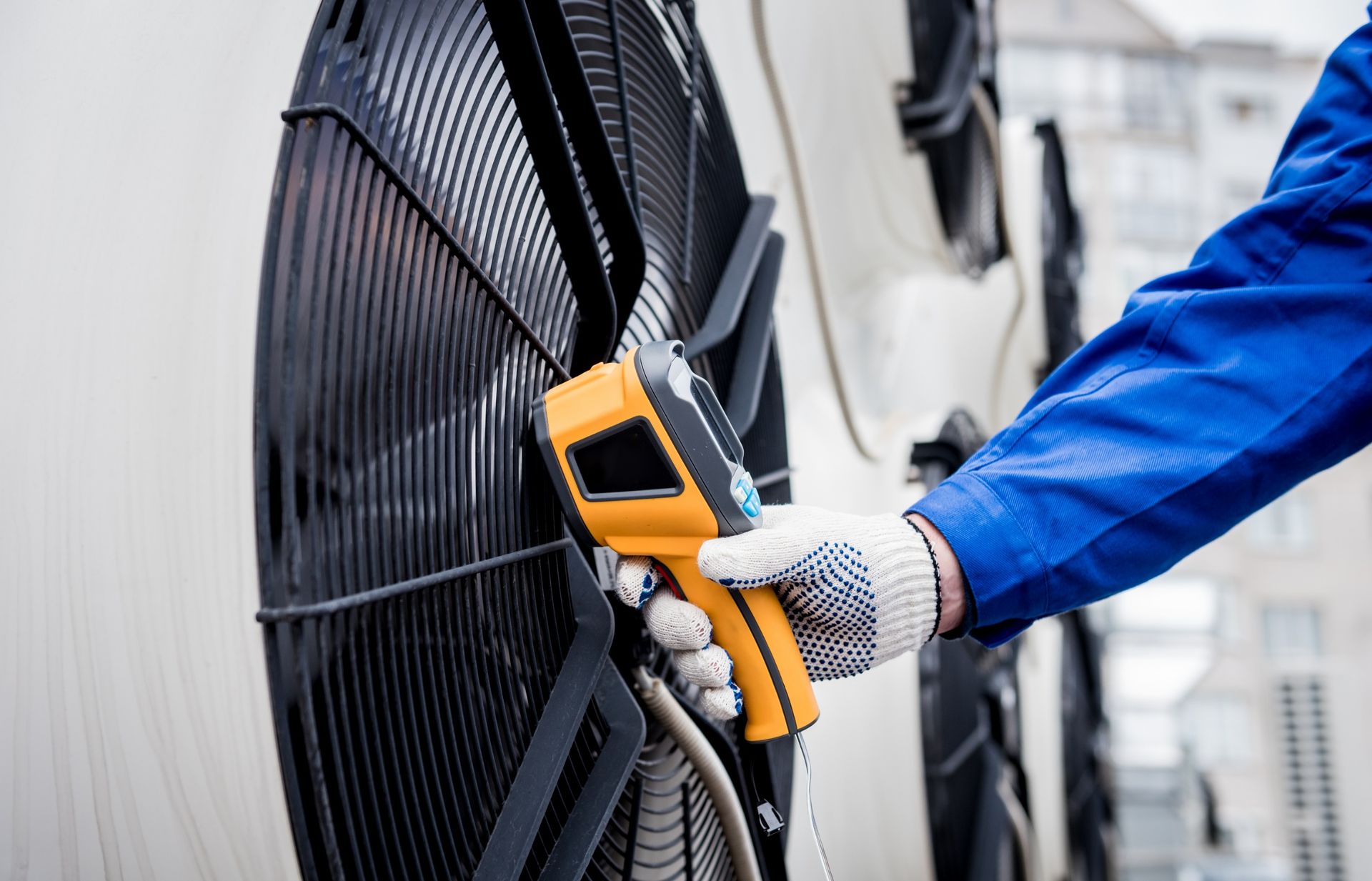 Person in blue coveralls uses a yellow infrared thermometer on an outdoor fan.